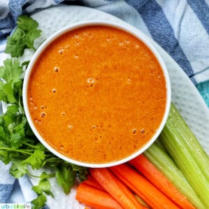 bowl of vegan buffalo sauce on a plate with carrot and celery sticks and parsley garnish.