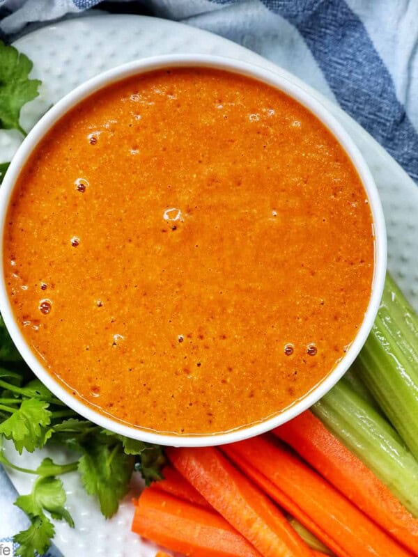 bowl of vegan buffalo sauce on a plate with carrot and celery sticks and parsley garnish.