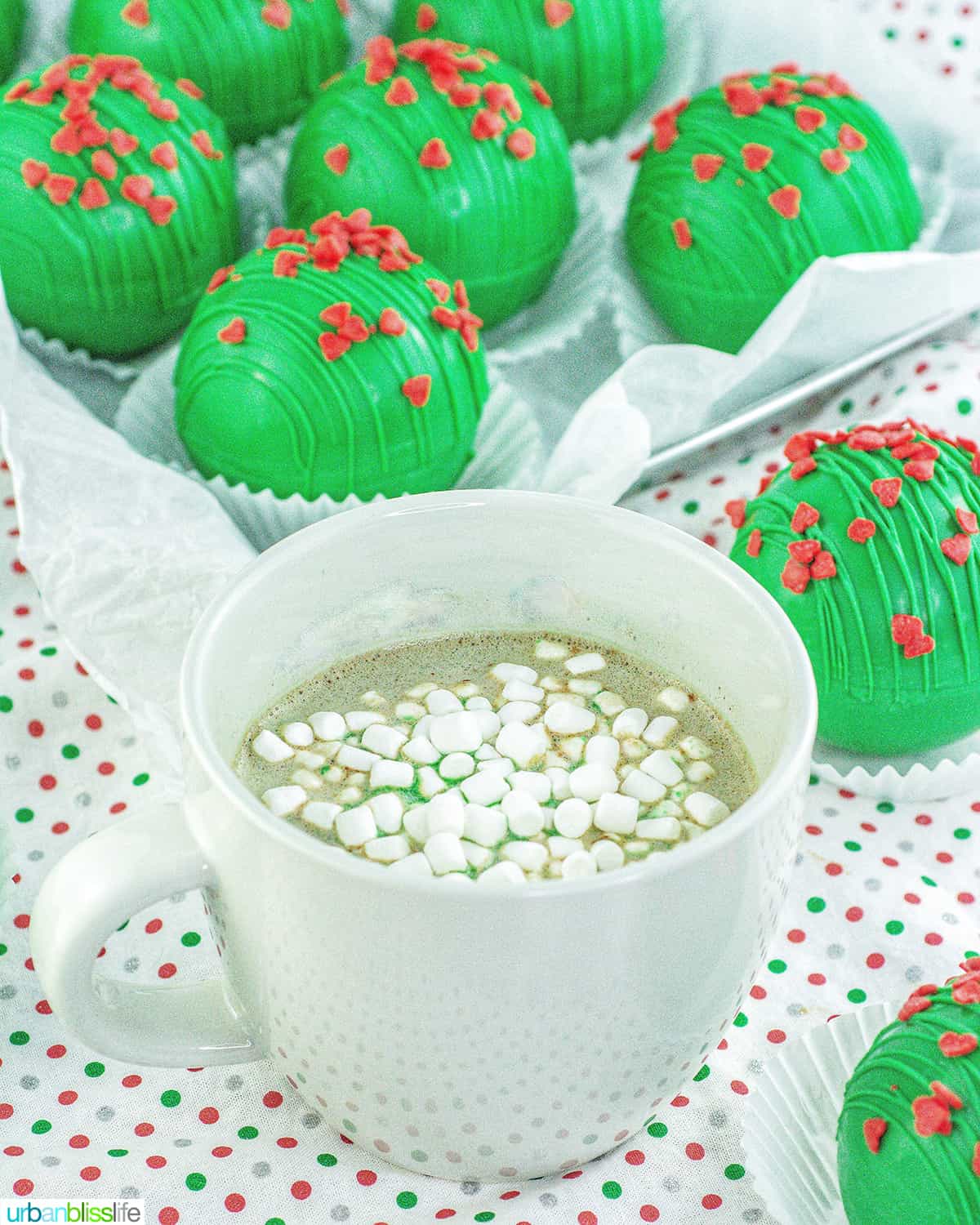 Grinch hot chocolate bombs around a mug of hot cocoa with marshmallows.