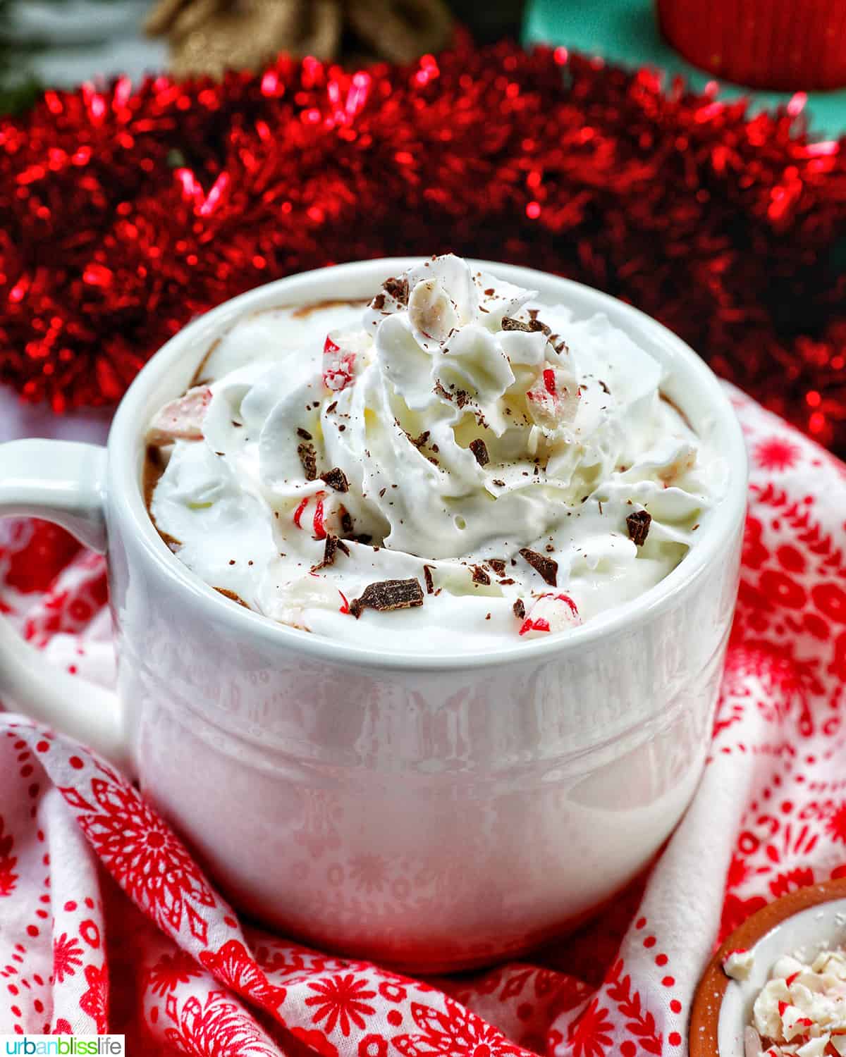 white mug with peppermint hot chocolate, whipped cream, surrounded by red and white Christmas decorations.