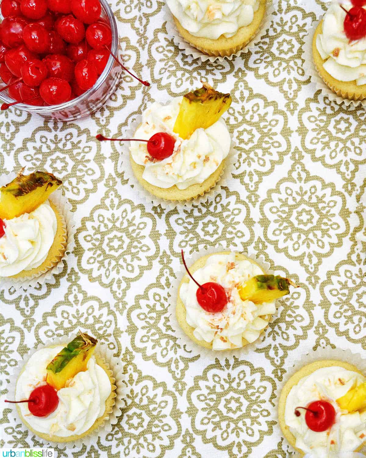 pina colada cupcakes on a patterned table with bowl of maraschino cherries.
