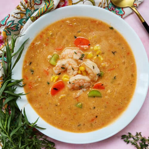 bowl of shrimp bisque with shrimp, corn, and carrots surrounded by fresh herbs.