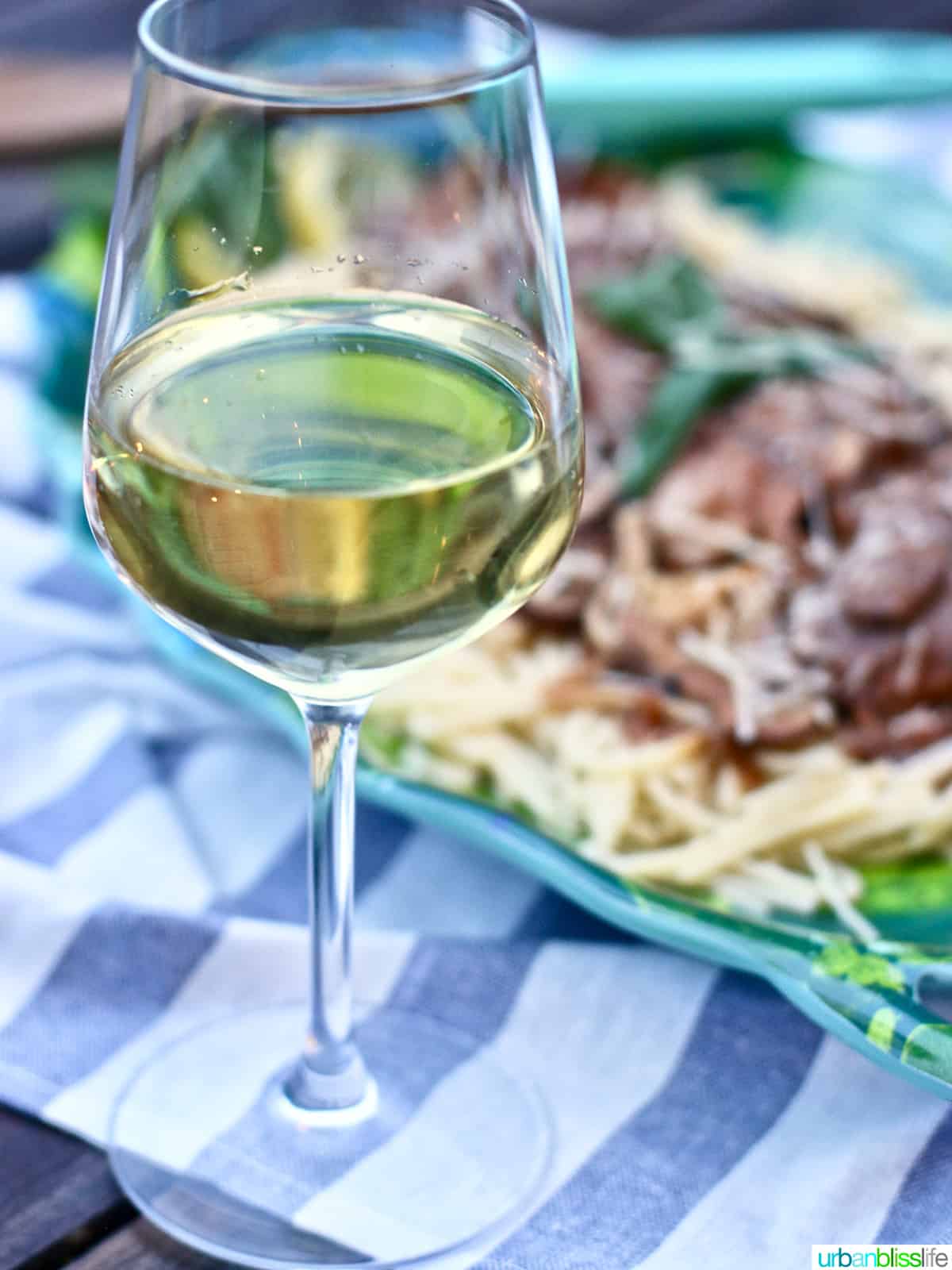 glass of white wine with pasta and meat dish behind.