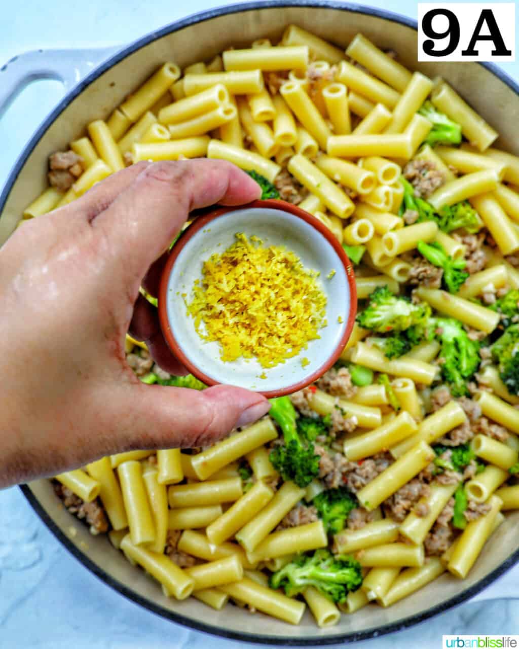 hand holding bowl of lemon zest over a skillet of broccoli and Italian sausage.