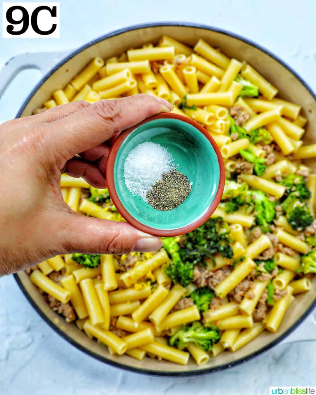 hand holding bowl of salt and pepper over a skillet of broccoli and Italian sausage.