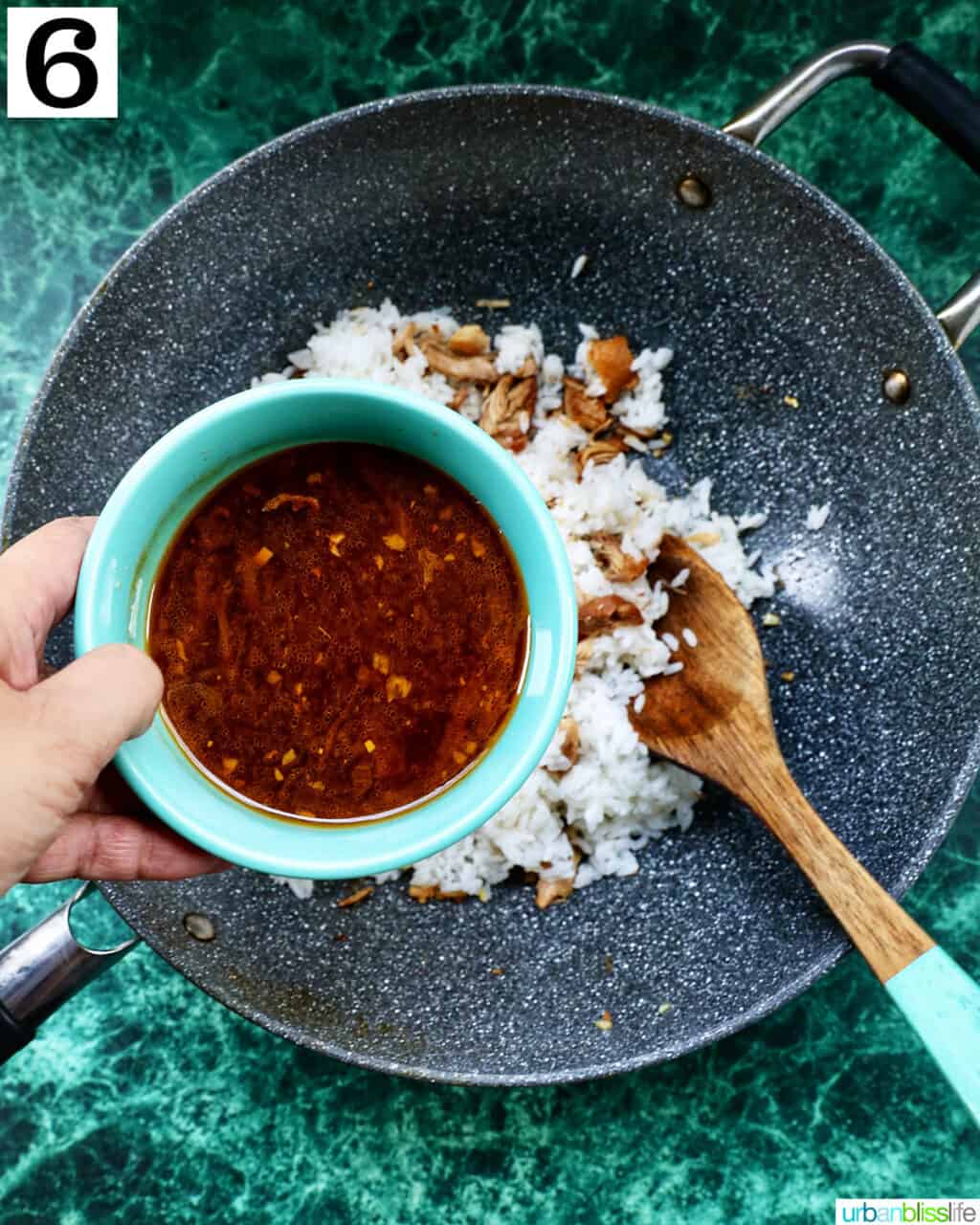 hand holding adobo sauce over a wok of adobo chicken and rice and a wooden spoon.