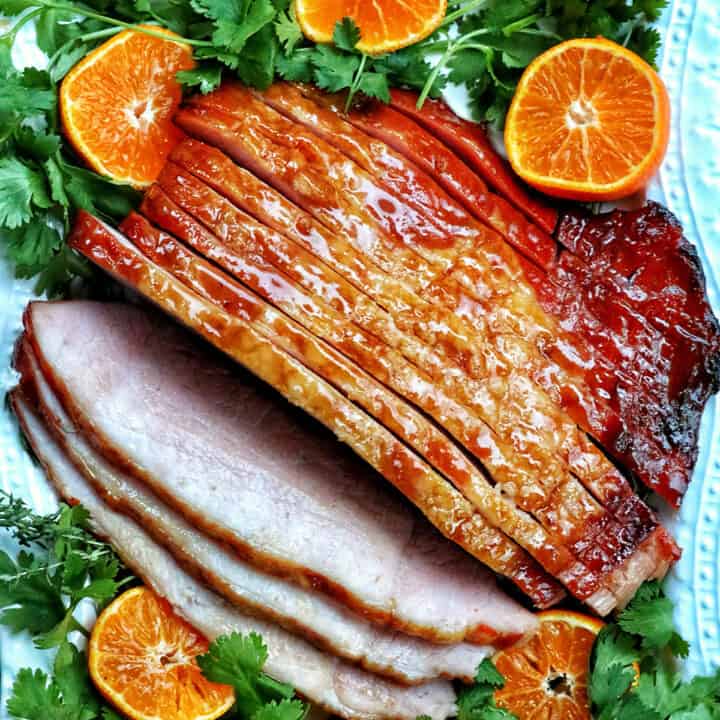 sliced honey glazed air fryer ham on a plate with fresh herbs and sliced oranges.