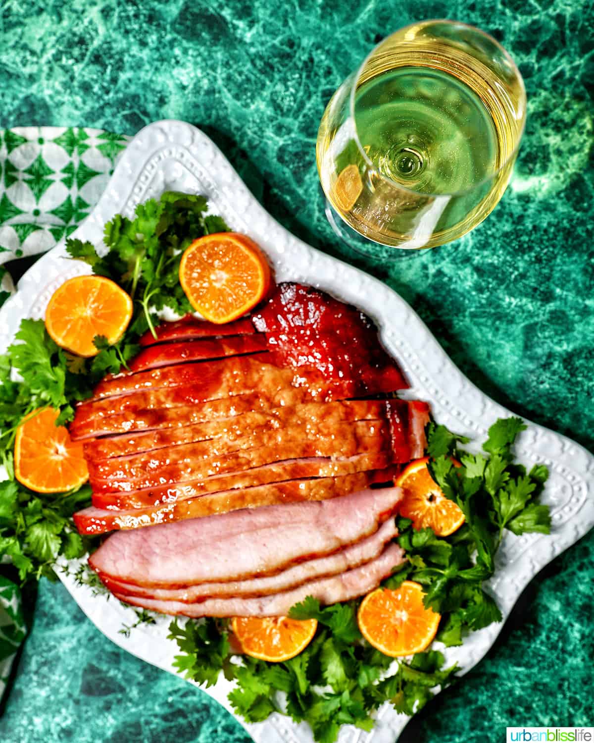 Glass of white wine behind a platter of honey glazed ham with garnishes.