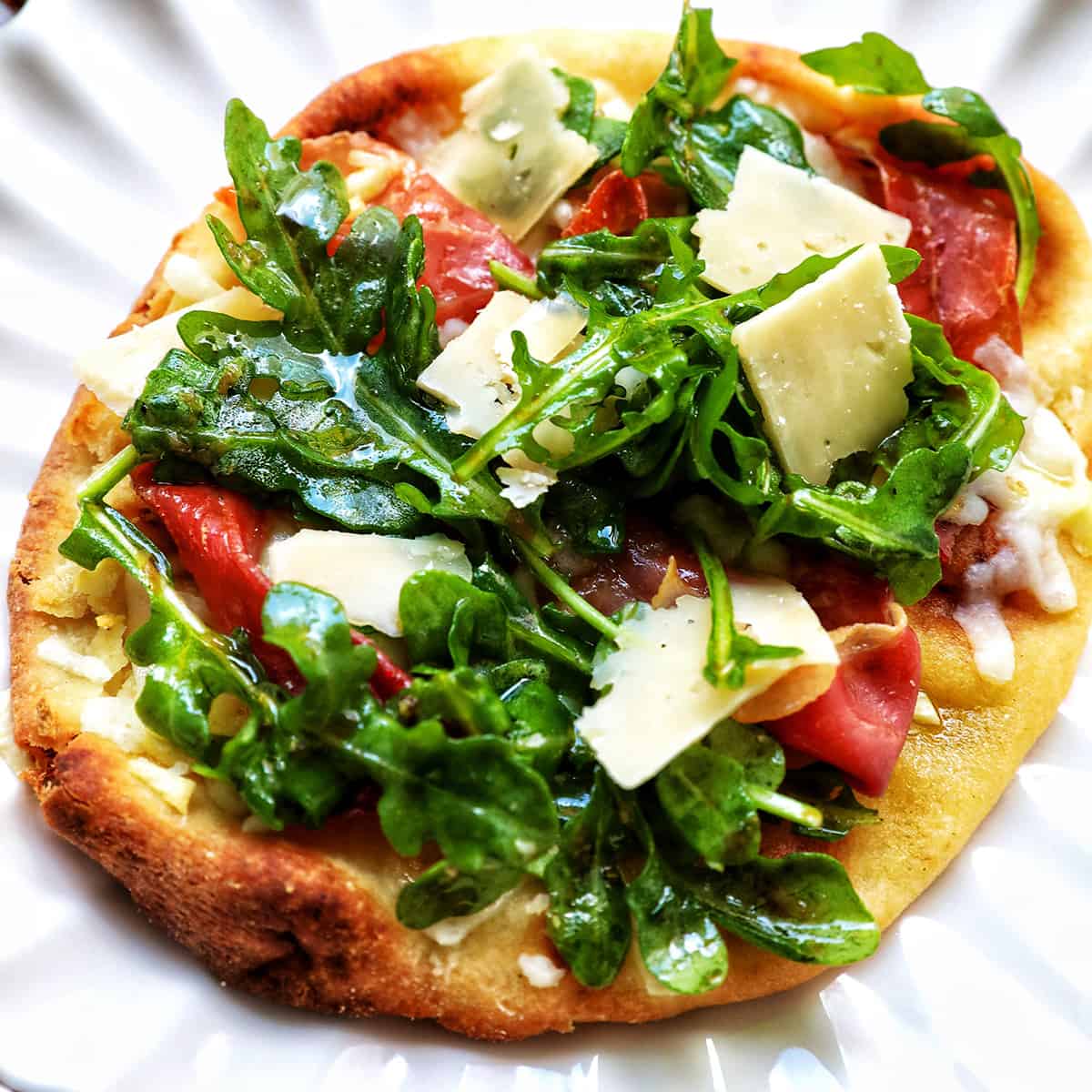 prosciutto flatbread with arugula and parmesan cheese on a scalloped plate.
