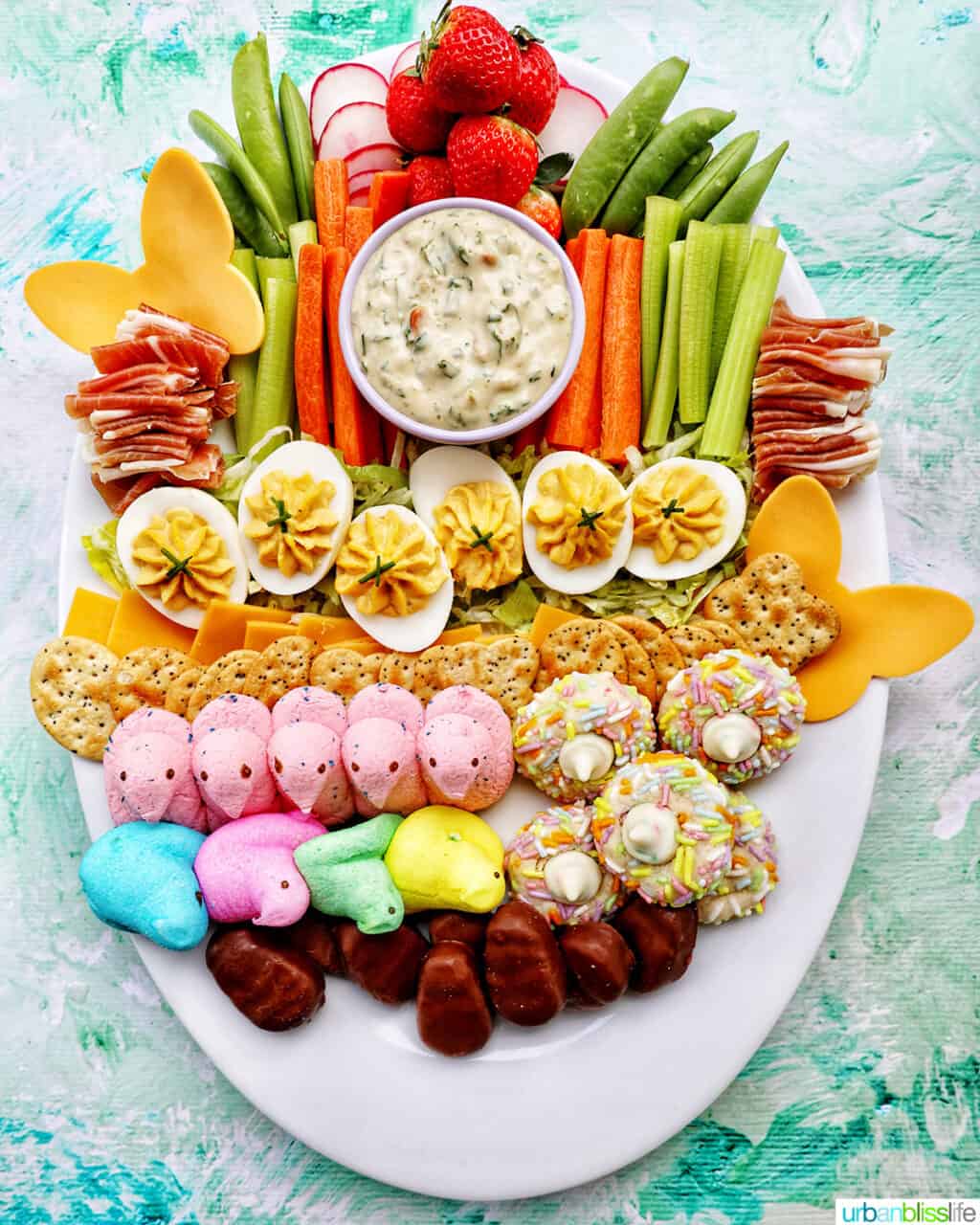 Peeps, chocolate Easter candies, crackers, and deviled eggs on shredded lettuce next to sliced vegetables and a bowl of vegetable dip on a large white platter.