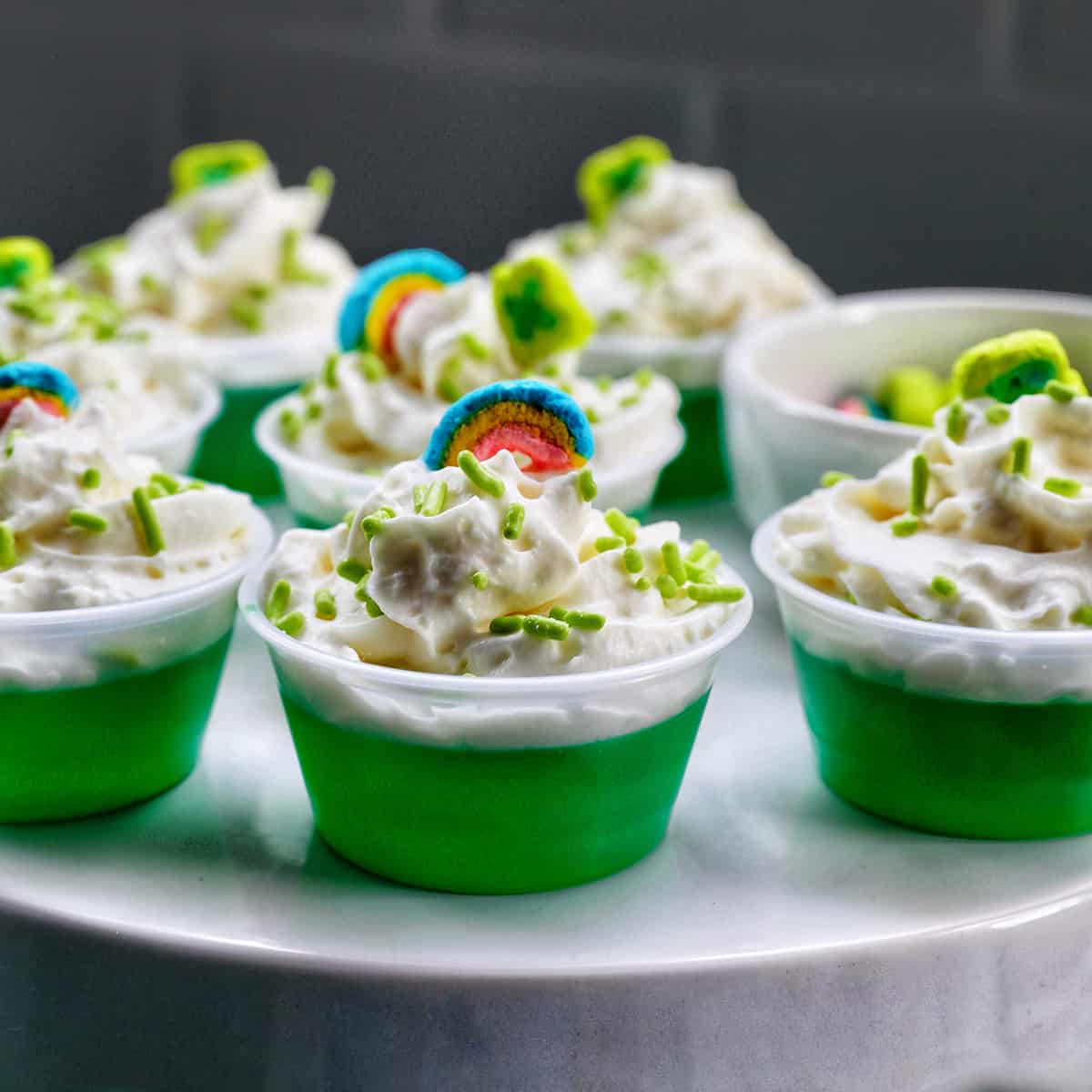 several green jello shots topped with whipped cream and colorful marshmallows on a cake pedestal.