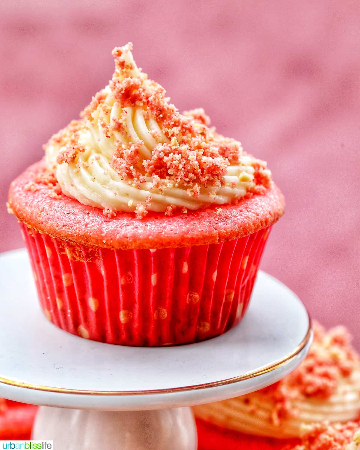 A strawberry cupcake with vanilla frosting and strawberry crunch topping on a white cupcake pedestal.