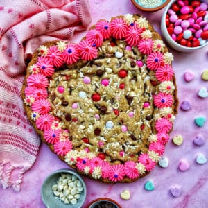 A Valentine's Day Cookie Cake with pink frosting, pink, red and white M & M candies, conversation hearts candies sprinkled around the cookie cake, and white sprinkles.