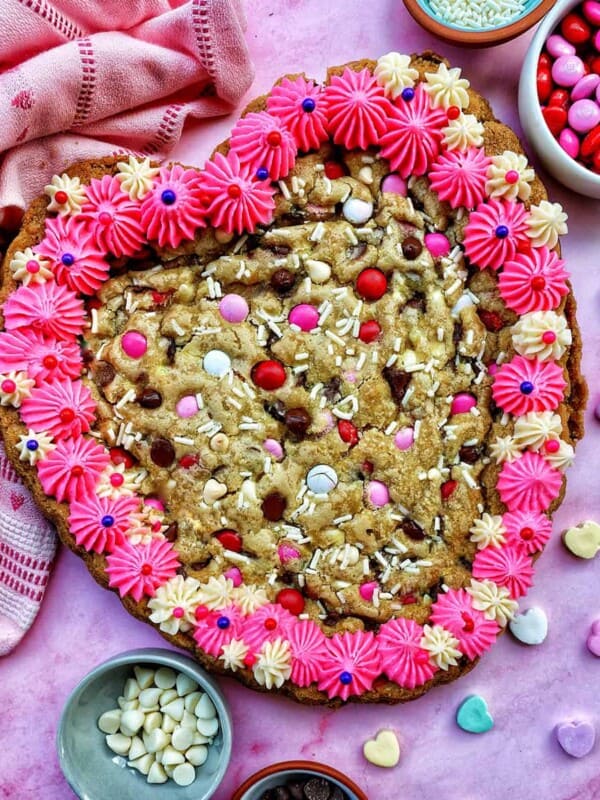 A Valentine's Day Cookie Cake with pink frosting, pink, red and white M & M candies, conversation hearts candies sprinkled around the cookie cake, and white sprinkles.