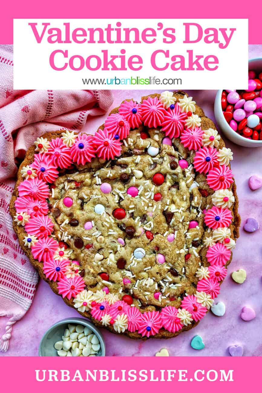 A Valentine's Day Cookie Cake with pink frosting, pink, red and white M & M candies, conversation hearts candies sprinkled around the cookie cake, and white sprinkles.