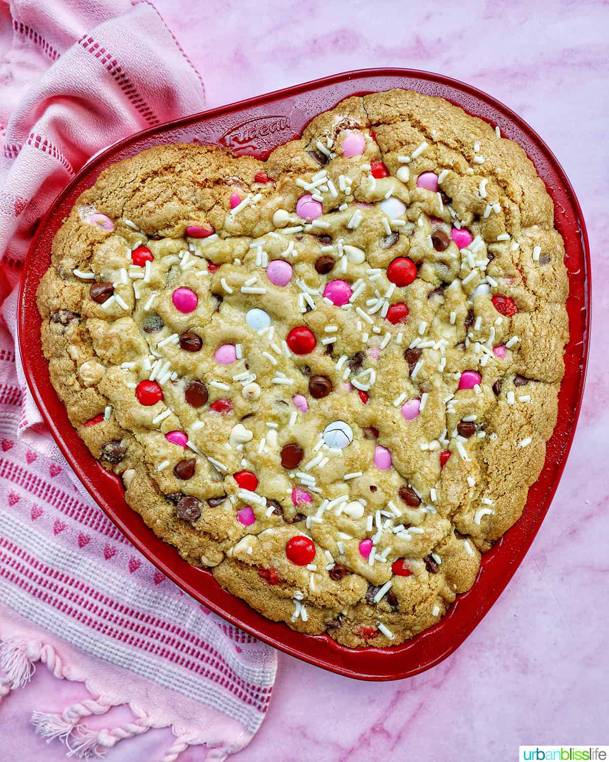 a heart-shaped cookie cake baked in a red pan on pink background.