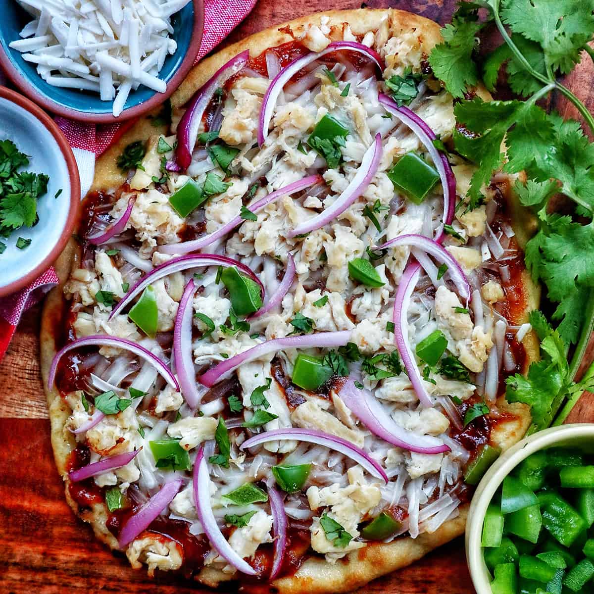 barbecue chicken flatbread pizza on a wooden board with sides of cheese and chopped cilantro for garnish.