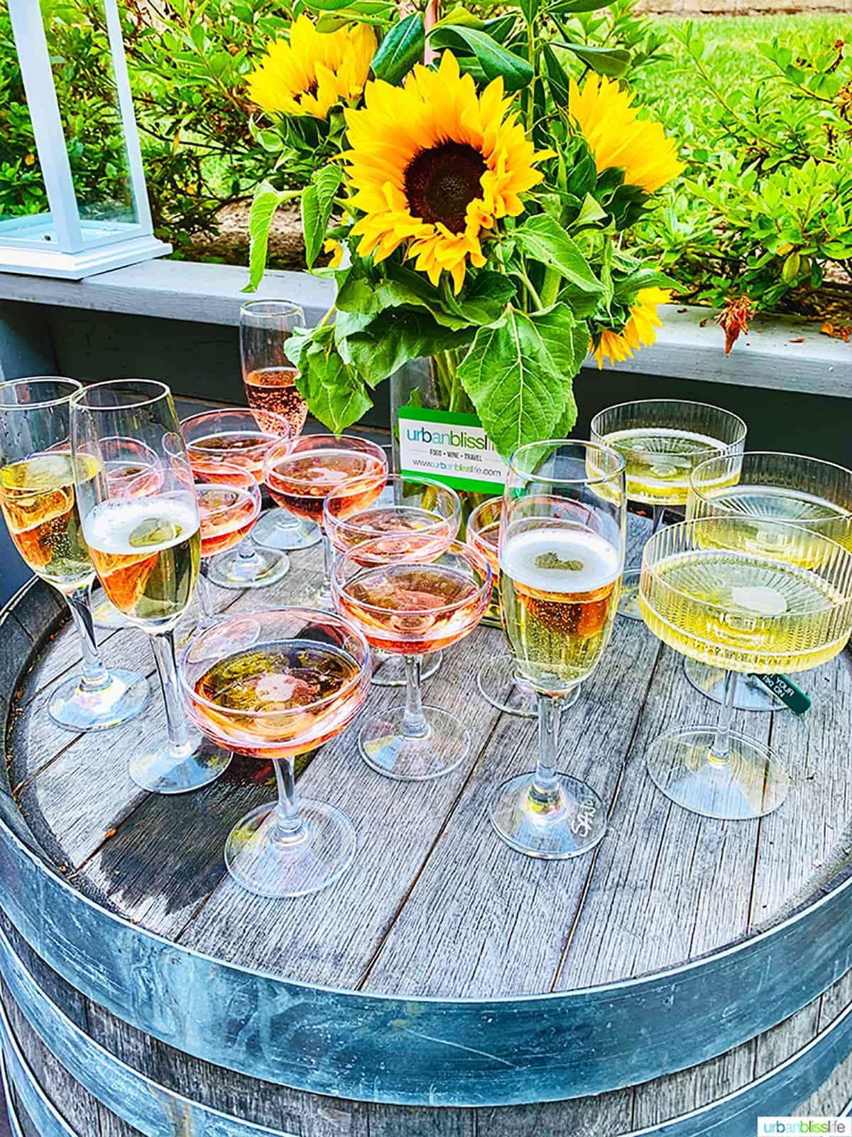 several glasses of rosé wine and Champagne on a wine barrel with vase of sunflowers.
