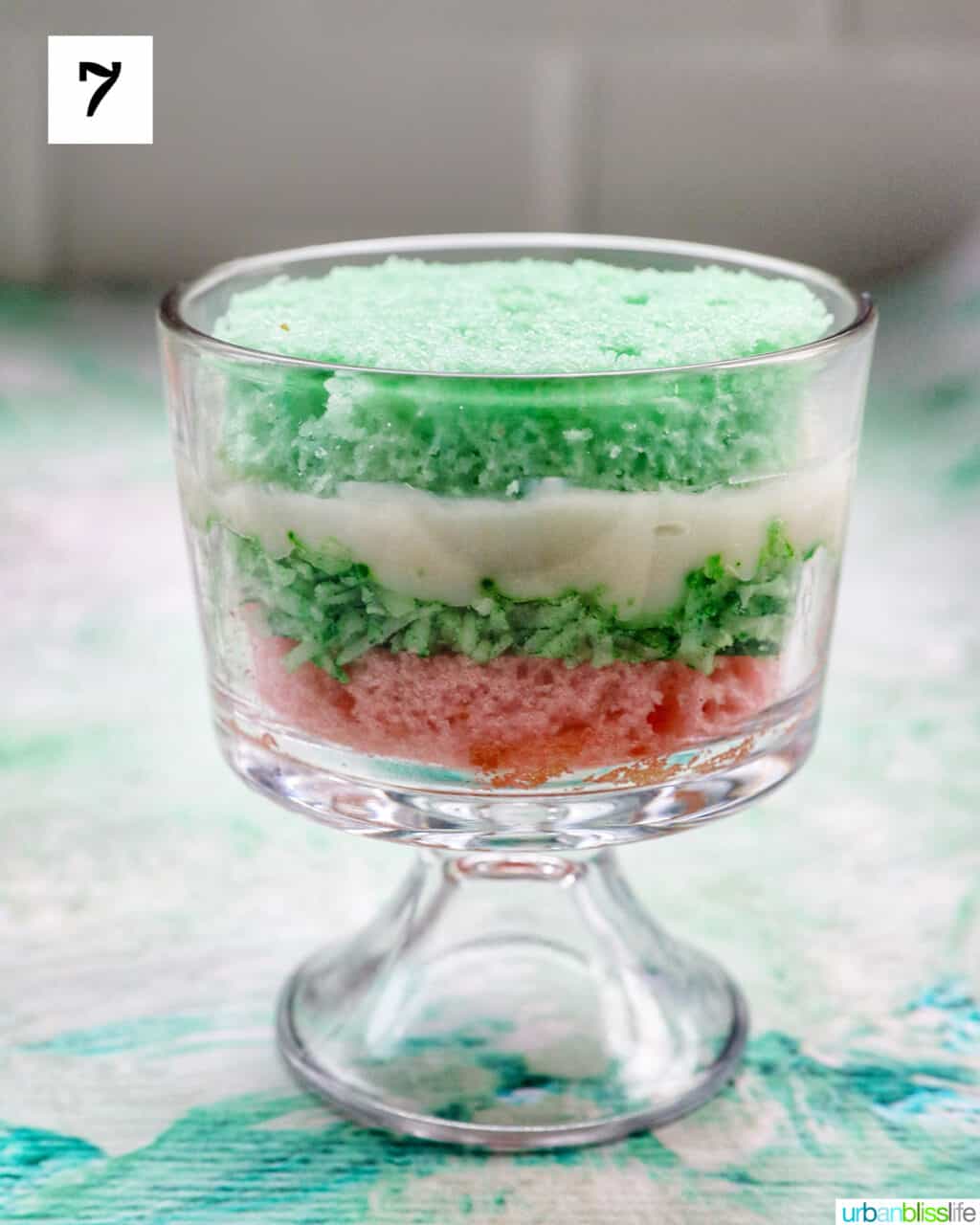 layer of green cake over vanilla pudding over shredded coconut over a layer of pink cake in a trifle bowl.