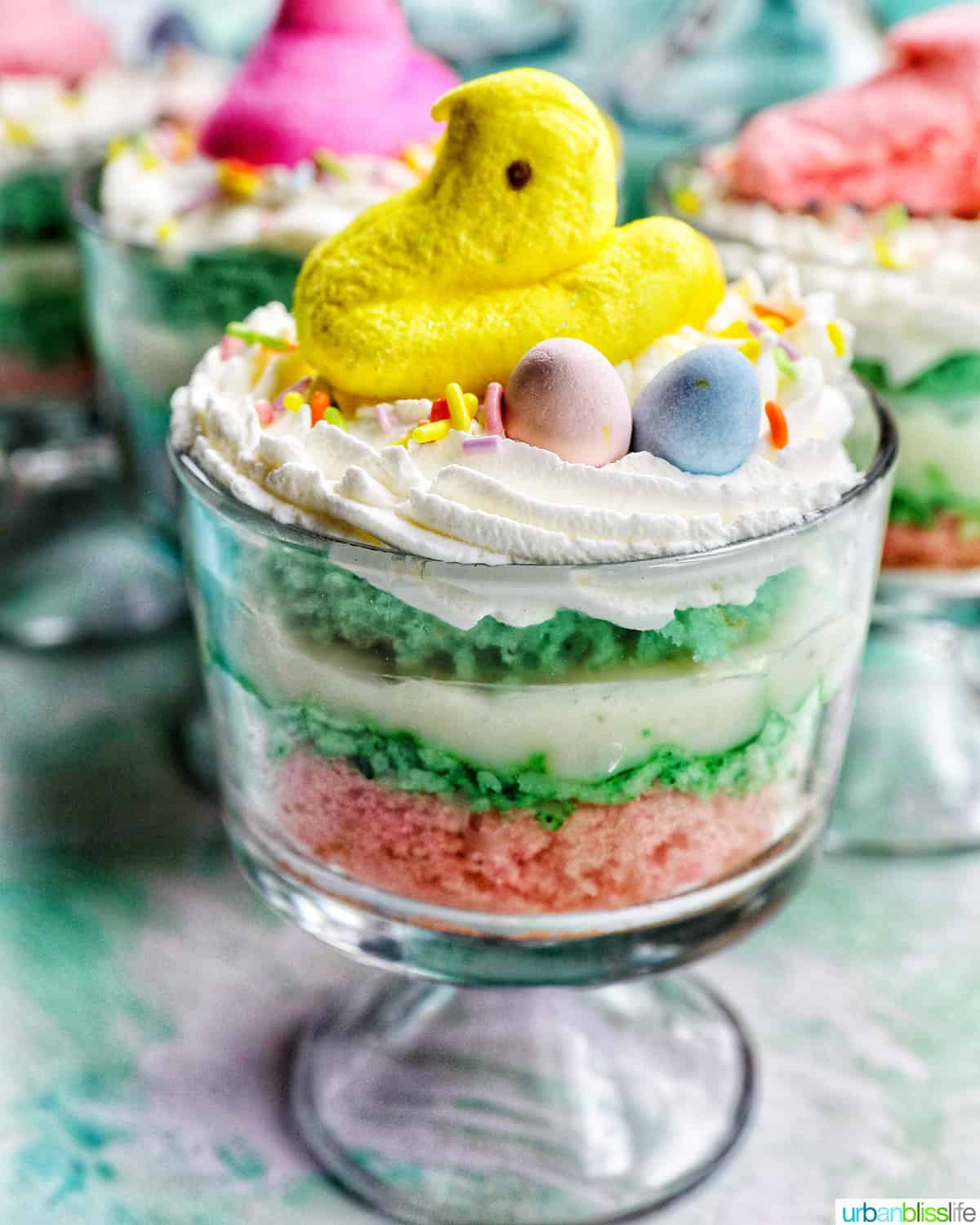 Easter trifle with layers of pastel colored cake, shredded coconut, vanilla pudding, and whipped cream, topped with colorful Easter candies.