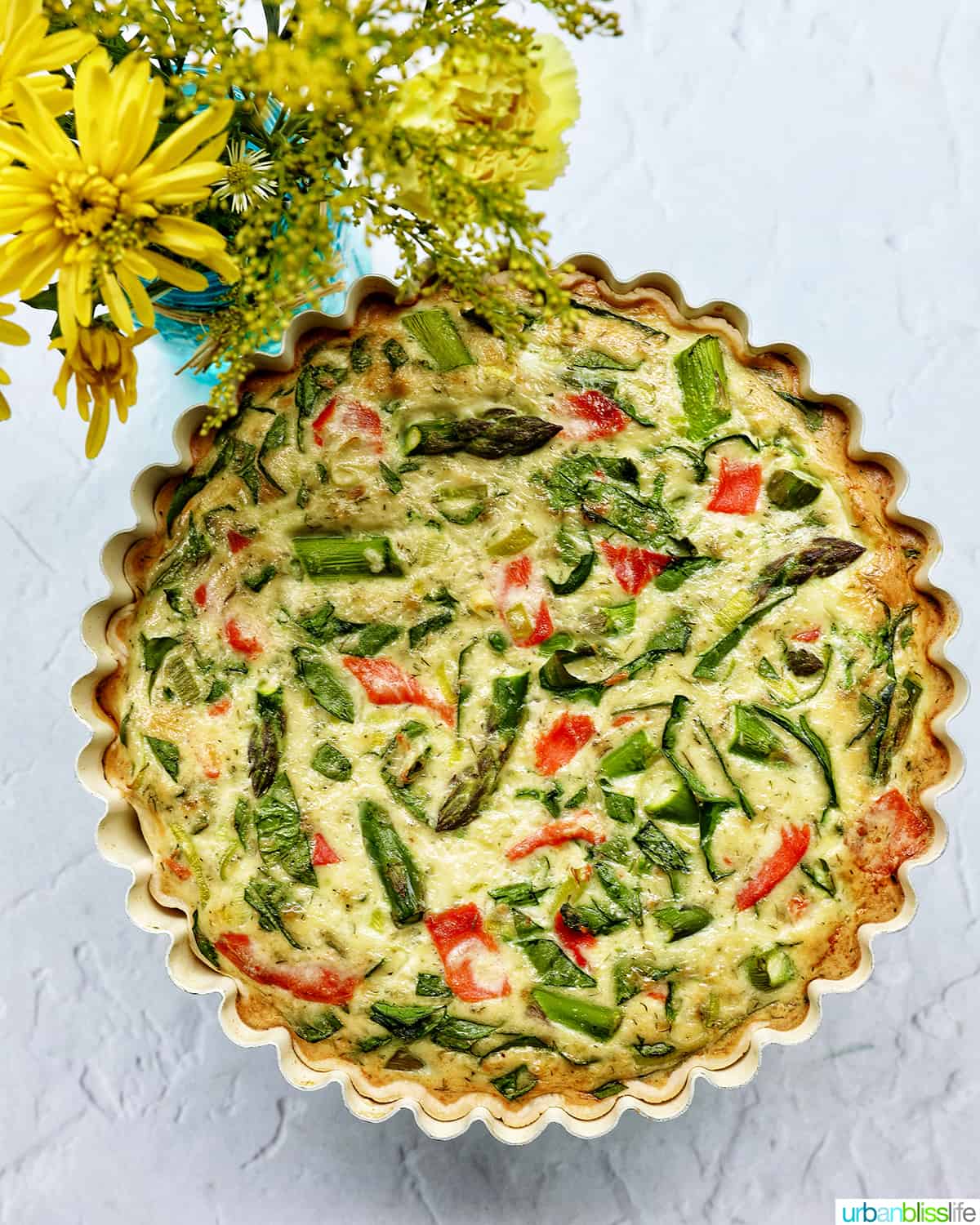 A quiche in a pie pan with salmon, asparagus, and leeks in front of a vase of yellow flowers.