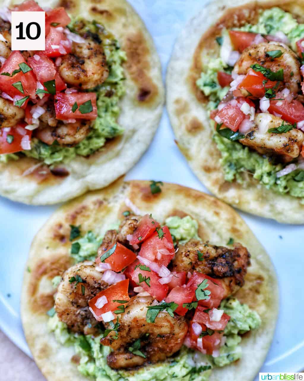 three shrimp tostafas with pico de gallo, shrimp, and guacamole on a tortilla with cilantro leaves on the side.