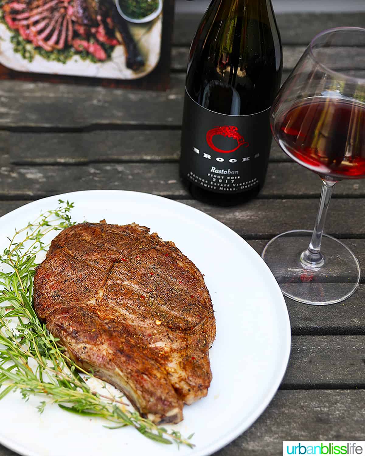 glass and bottle of red wine next to a plate of steak and herbs.