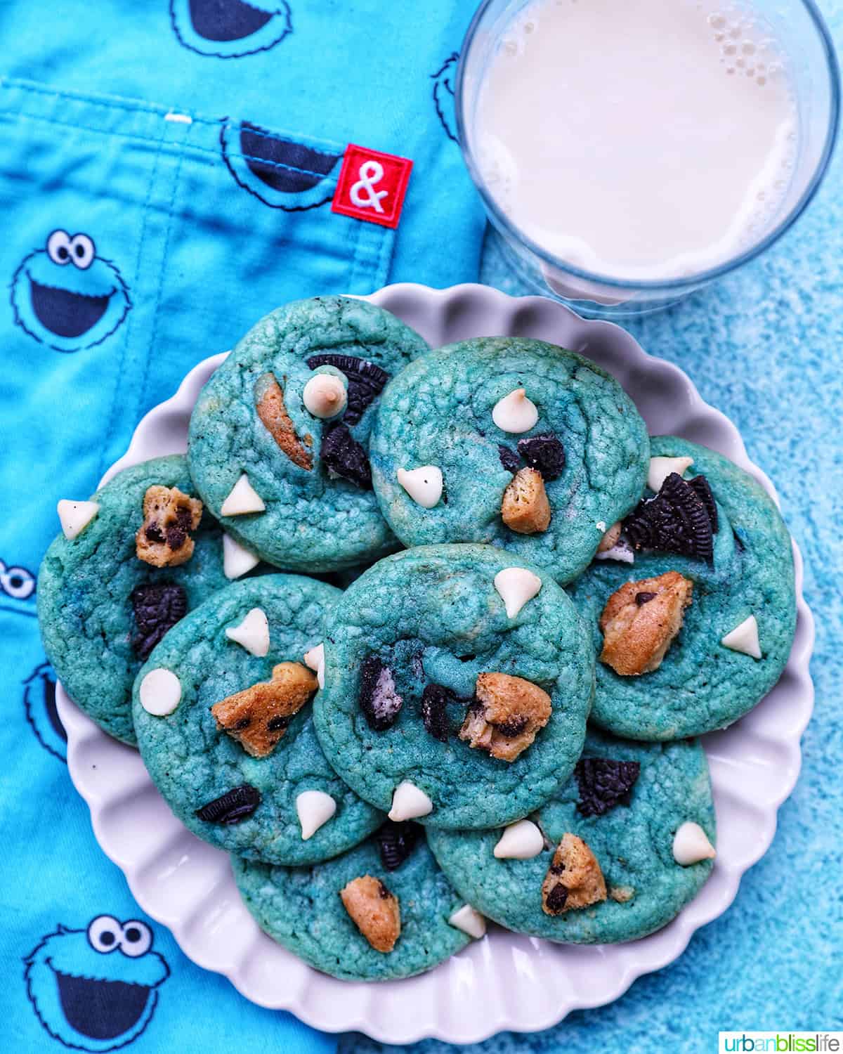 plate full of bright blue Cookie Monster Cookies on top of a blue Cookie Monster apron next to a glass of milk.