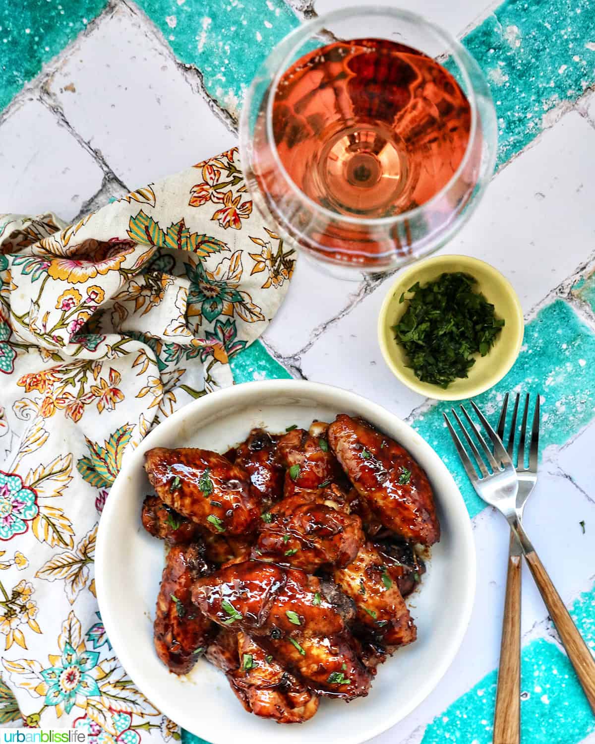 bowl of Filipino adobo chicken wings and fresh parsley, two forks, and a glass of rosé wine.