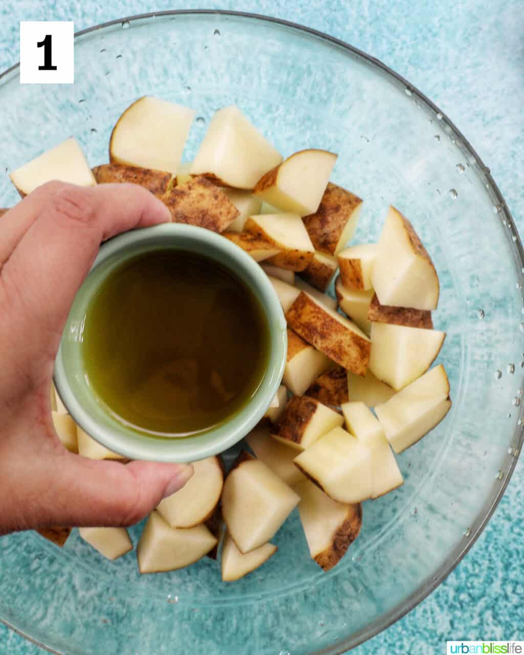 hand holding bowl of olive oil over a glass bowl with potato wedges.