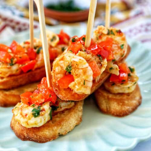 shrimp pintxos skewers with tomatoes on crostini bread on a scalloped plate.