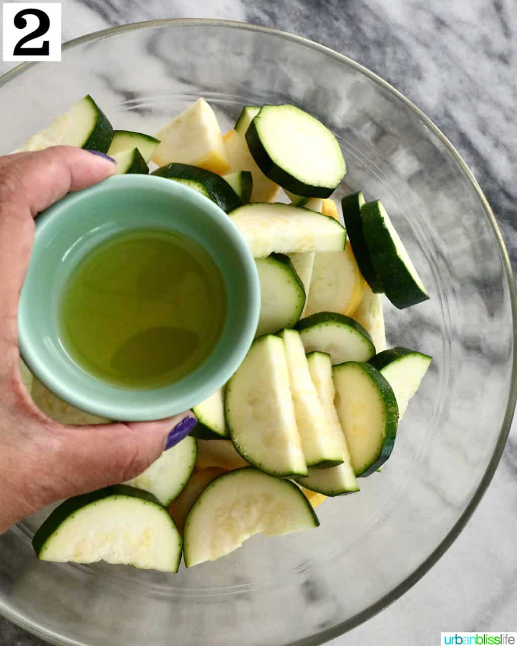 hand holding small bowl of olive oil over zucchini and yellow squash in a bowl.