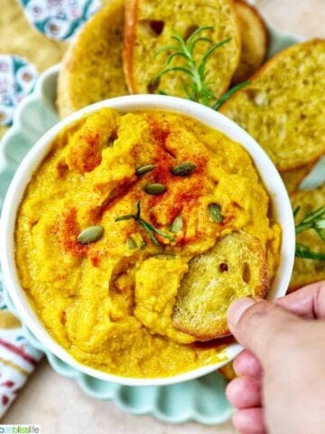 hand dipping crostini into bowl of butternut squash dip with baguette slices, fresh herbs, and a colorful napkin.