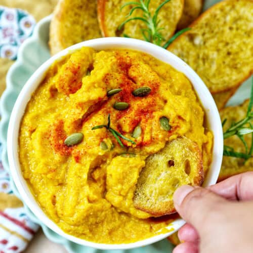 hand dipping crostini into bowl of butternut squash dip with baguette slices, fresh herbs, and a colorful napkin.