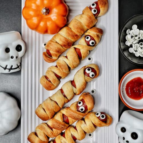 mummy hot dogs on a white plate surrounded by Halloween decorations like mini pumpkins and skull bowls.