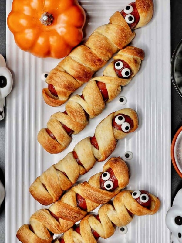 mummy hot dogs on a white plate surrounded by Halloween decorations like mini pumpkins and skull bowls.