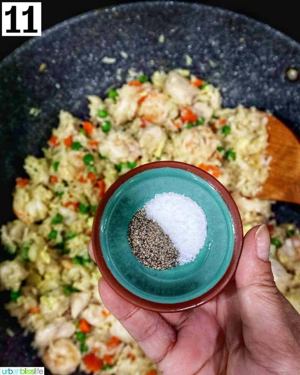 hand holding a small bowl of salt and pepper over a wok of shrimp and chicken fried rice.