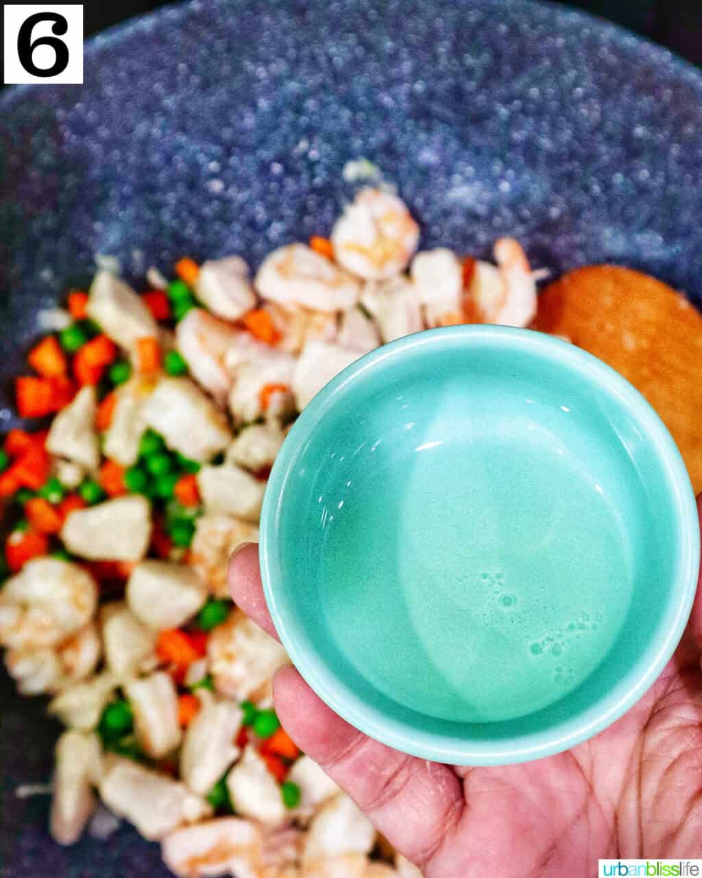 hand holding a bowl of oil over a wok with chicken and veggies.