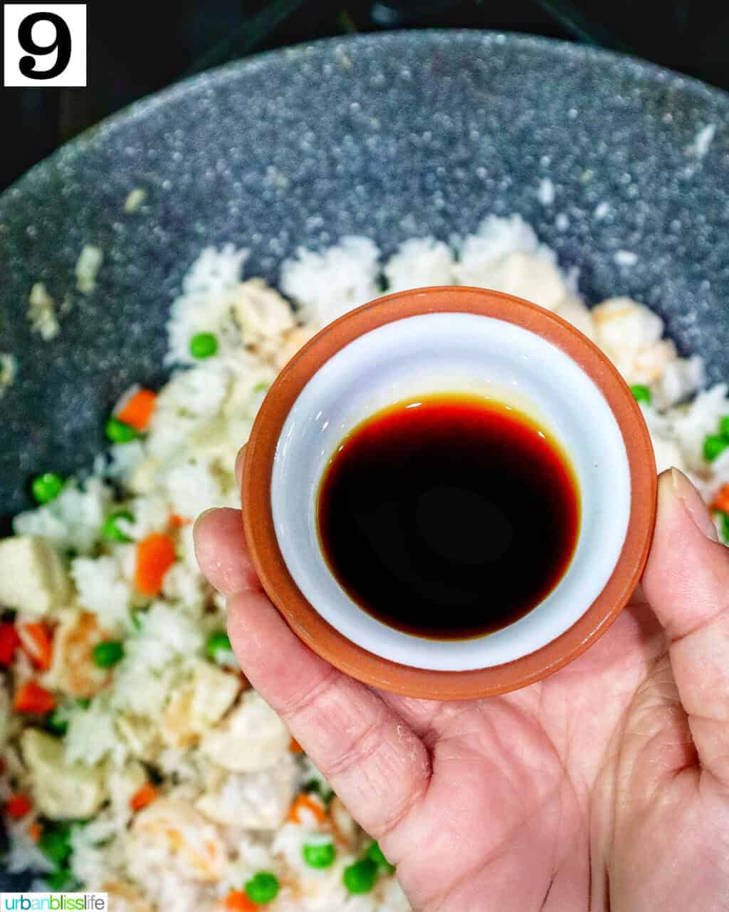 hand holding a small bowl of soy sauce over a wok of shrimp and chicken fried rice.