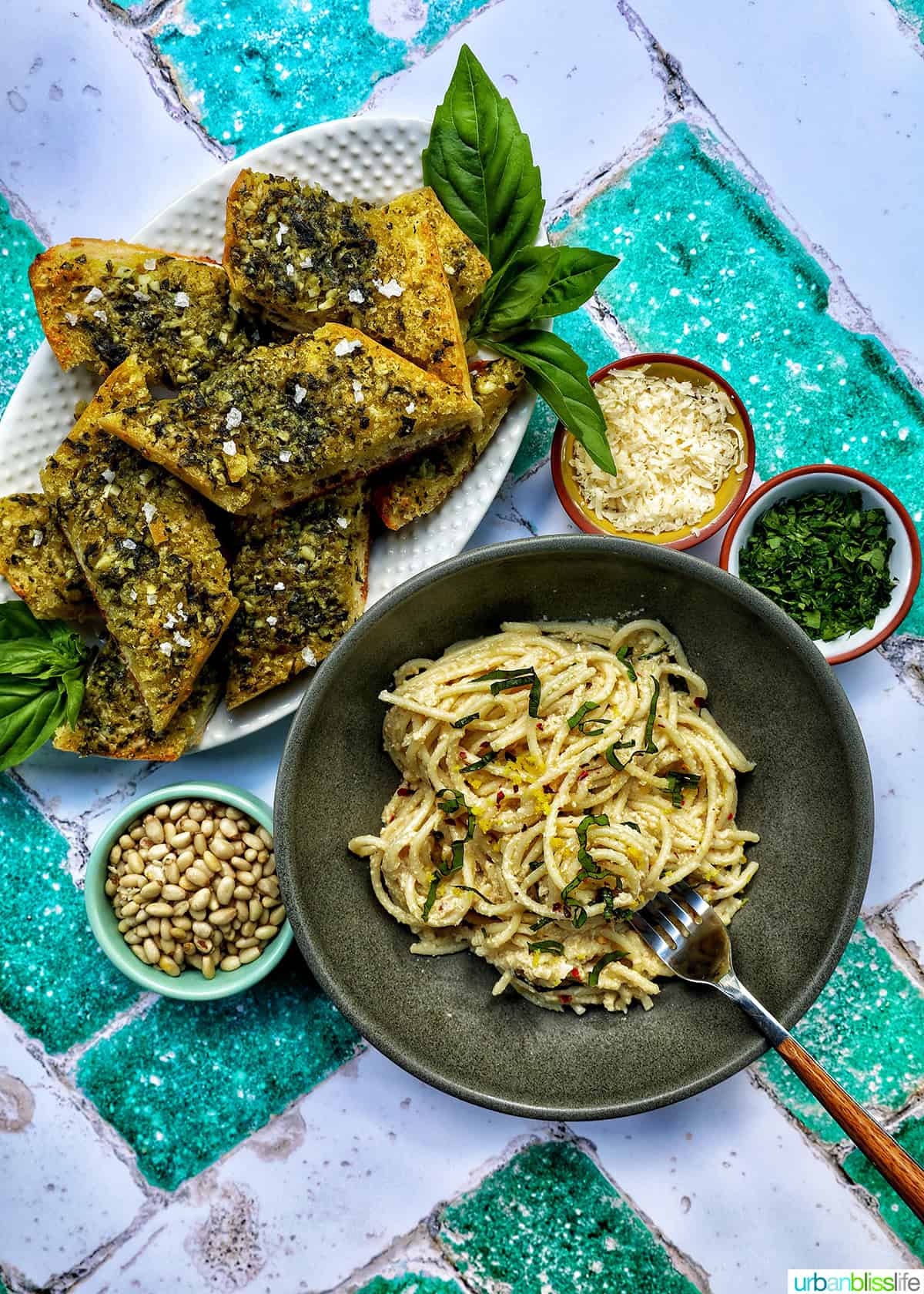 slices of pesto bread on a white platter with glass of white wine and bowl of tahini pasta, with bowls of garnishes.