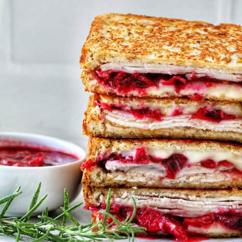 stack of Turkey Cranberry Brie Sandwiches sliced in half with side of cranberry sauce and herbs.