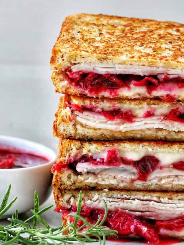 stack of Turkey Cranberry Brie Sandwiches sliced in half with side of cranberry sauce and herbs.