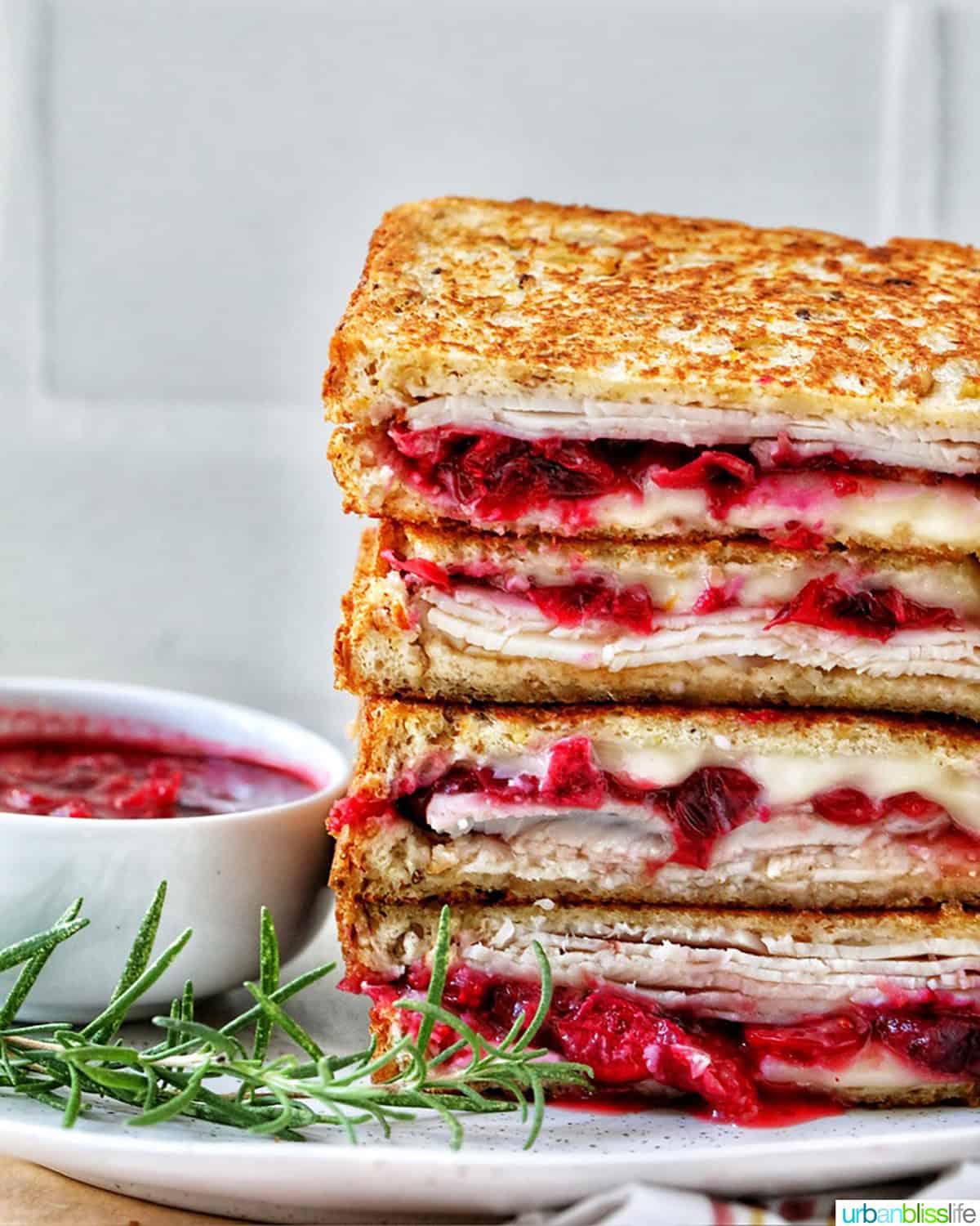 stack of Turkey Cranberry Brie Sandwiches sliced in half with side of cranberry sauce and herbs.