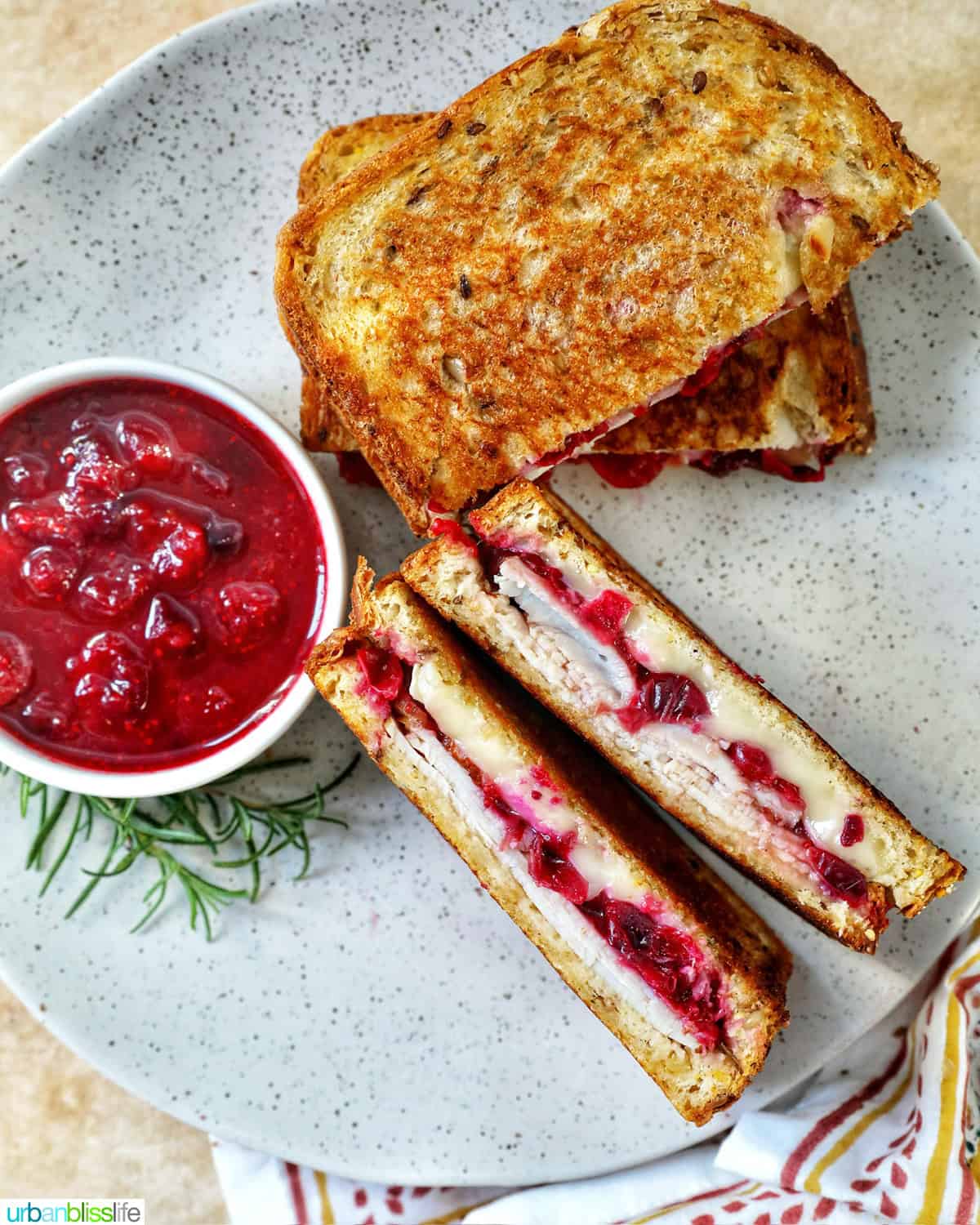 stack of Turkey Cranberry Brie Sandwiches sliced in half with side of cranberry sauce and herbs.