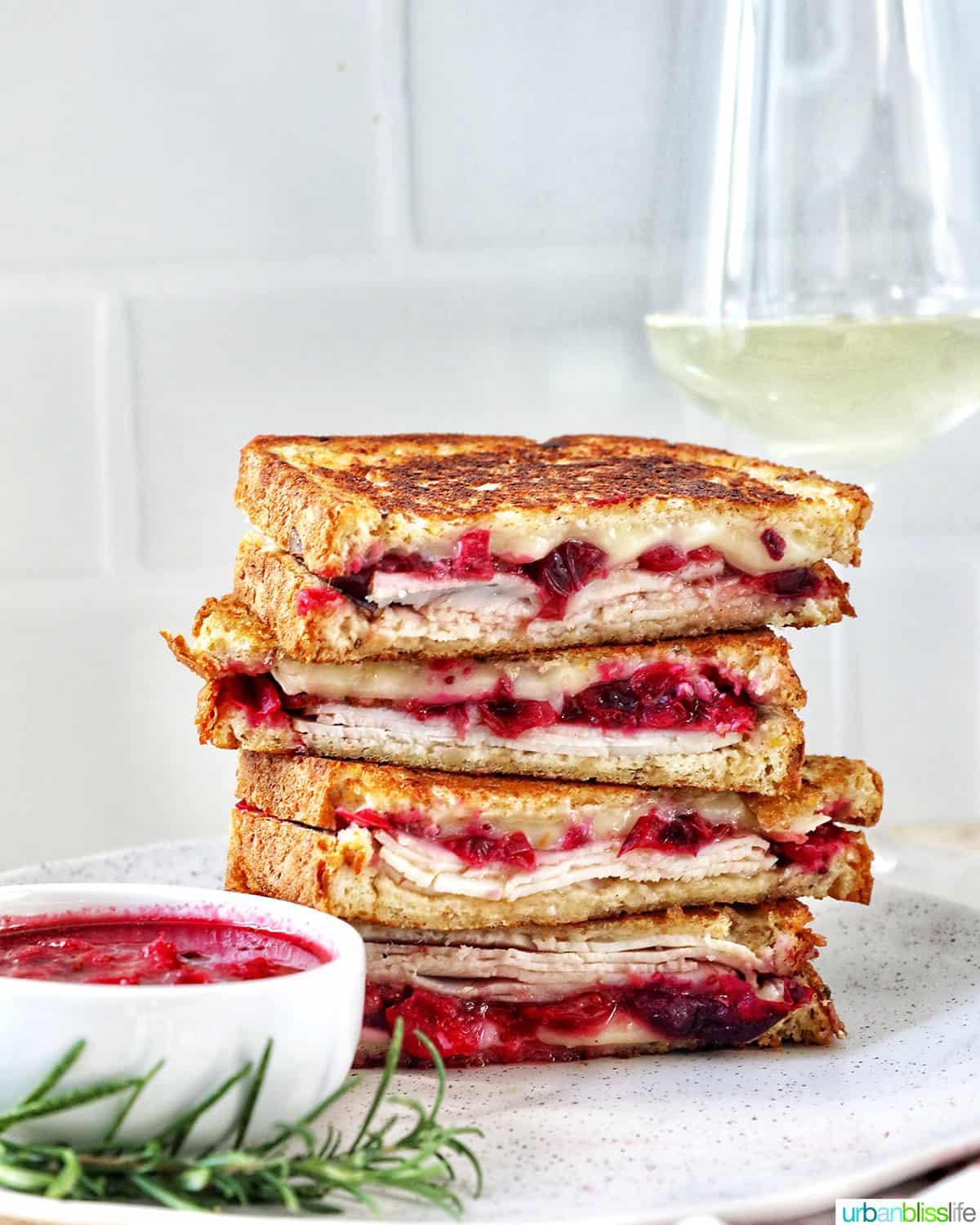 stack of Turkey Cranberry Brie Sandwiches sliced in half with side of cranberry sauce and herbs and glass of white wine.