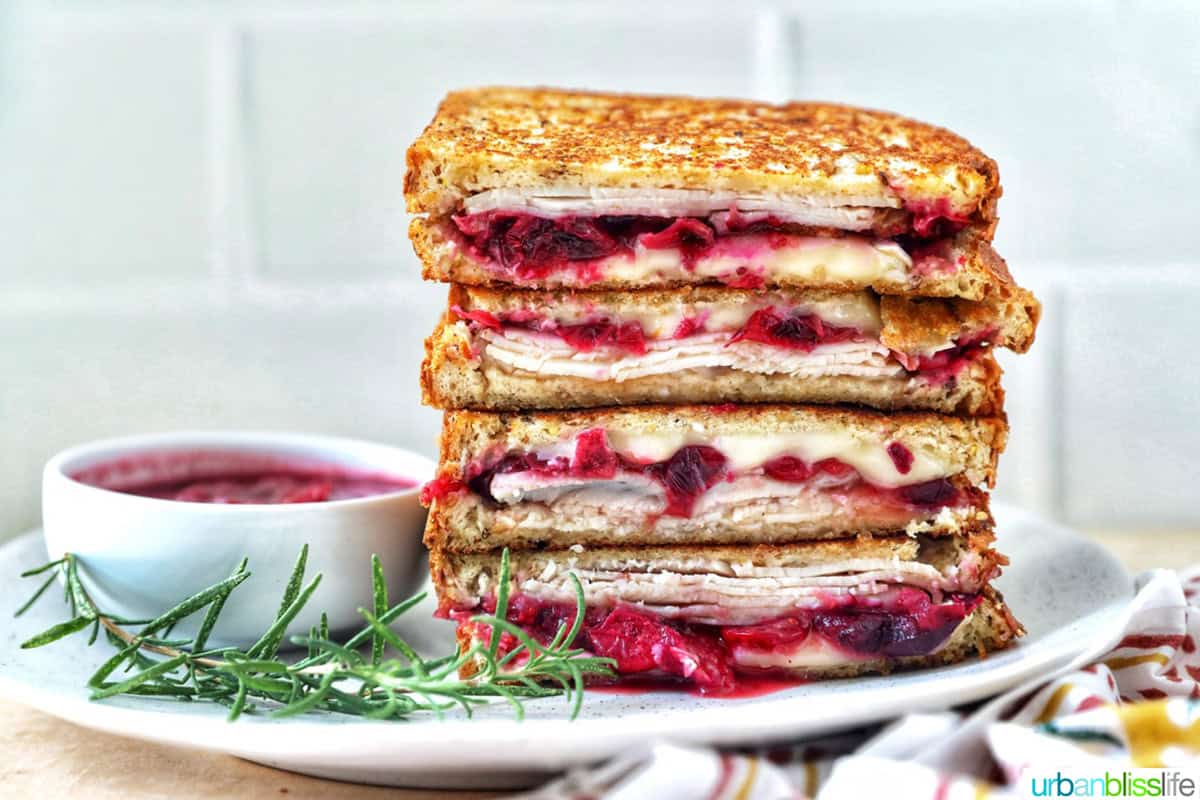 stack of Turkey Cranberry Brie Sandwiches sliced in half with side of cranberry sauce and herbs.