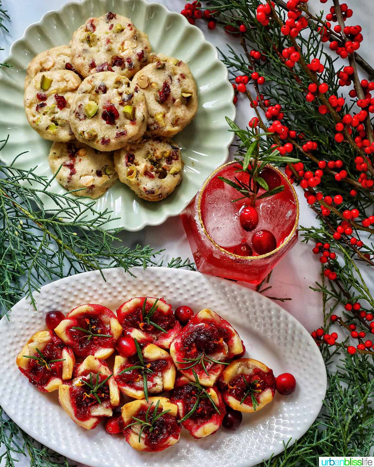 pistachio cranberry shortbread cookies piled onto a plate next to holiday berries and greenery, cranberry brie bites, a cranberry mocktail.