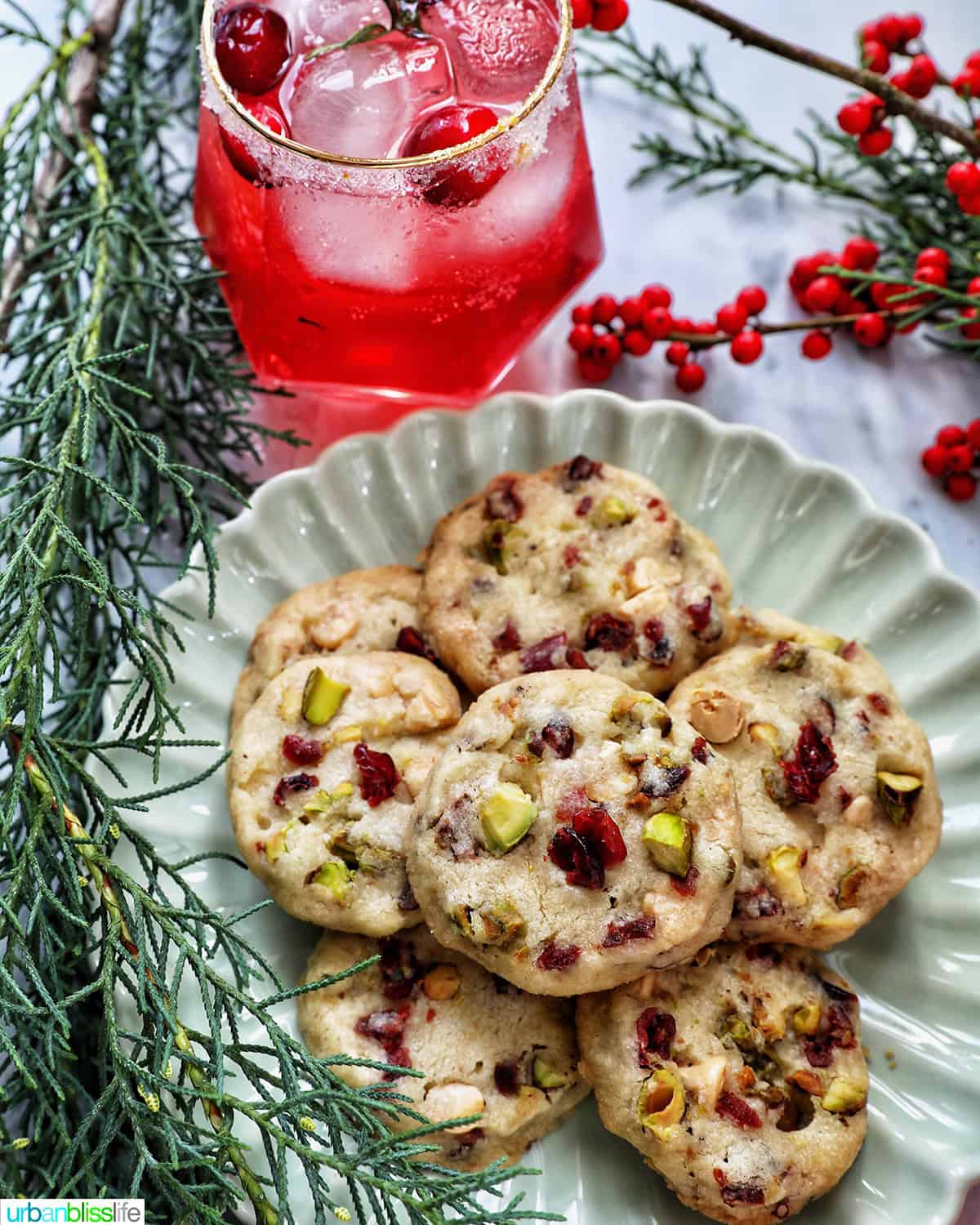 pistachio cranberry shortbread cookies piled onto a plate next to holiday berries and greenery and a cranberry mocktail.