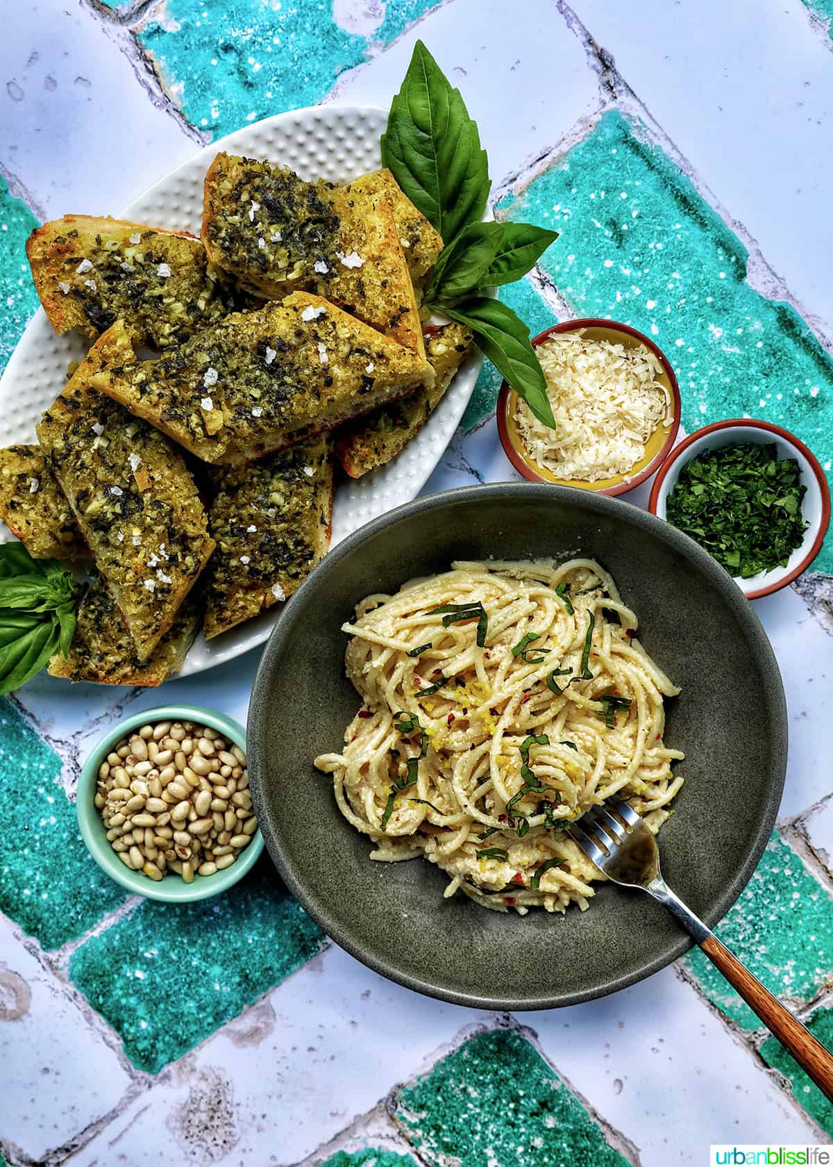 bowl of tahini pasta with plate of pesto bread and side bowls of pine nuts, cheese, and parsley.