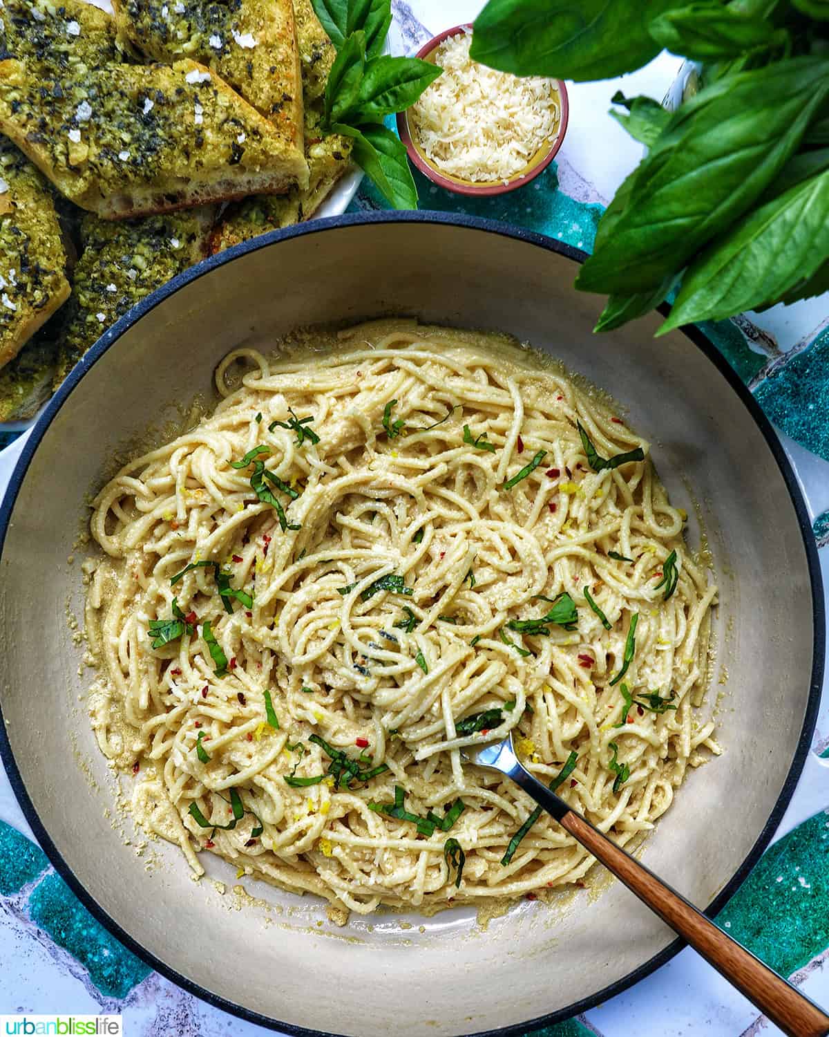 saucepan of tahini pasta with plate of pesto bread and side bowls of pine nuts, cheese, and parsley.